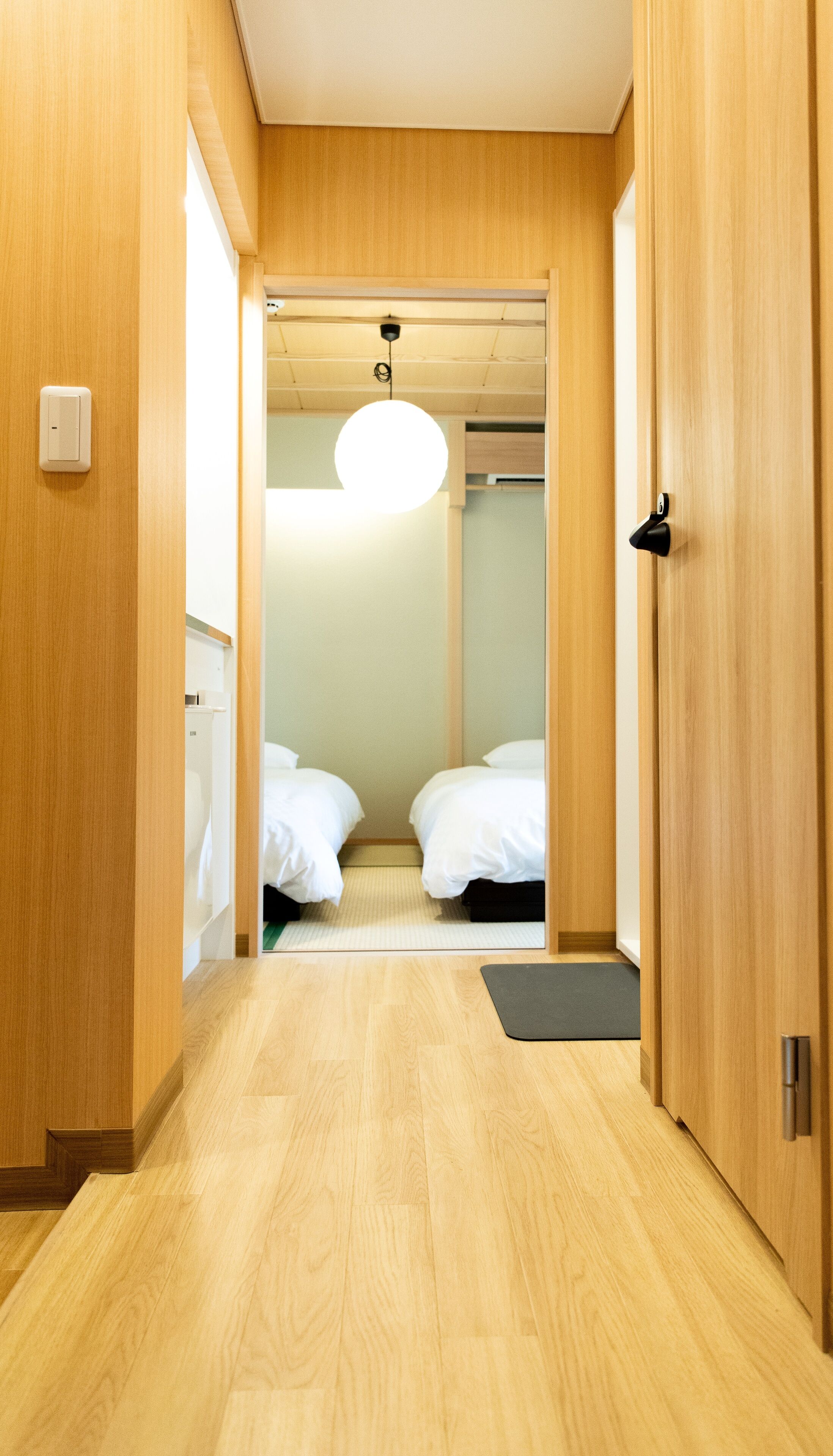 Un pasillo de hotel con piso de madera, una puerta que da a una habitación con dos camas, una lámpara en el techo y un felpudo pequeño en la entrada.