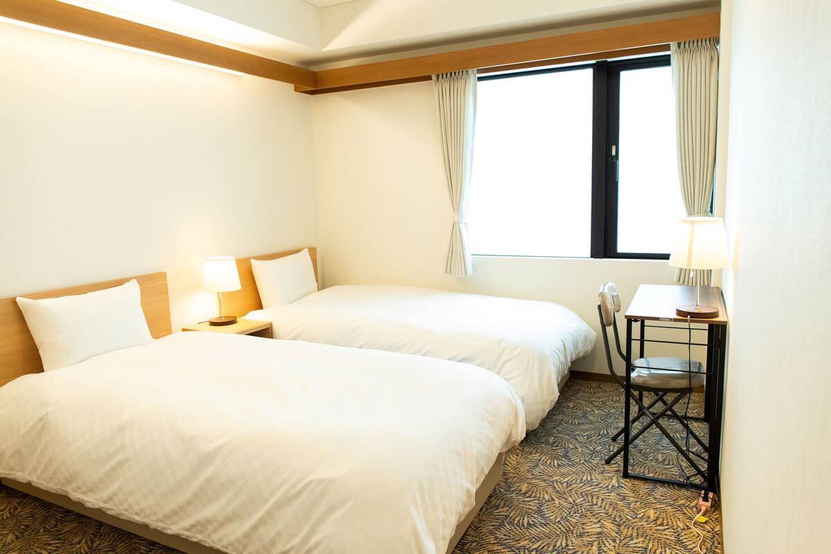 Une chambre d’hôtel avec deux lits, un bureau avec une chaise, une fenêtre avec des rideaux et une moquette à motifs.