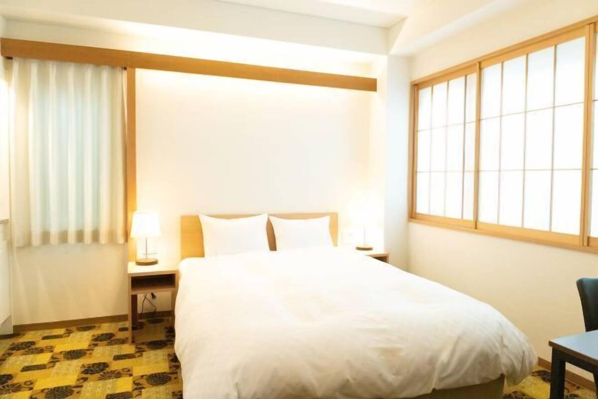 Une chambre d’hôtel comprenant un lit, une table de chevet, une lampe, une chaise, une fenêtre avec des stores et un tapis à motifs.