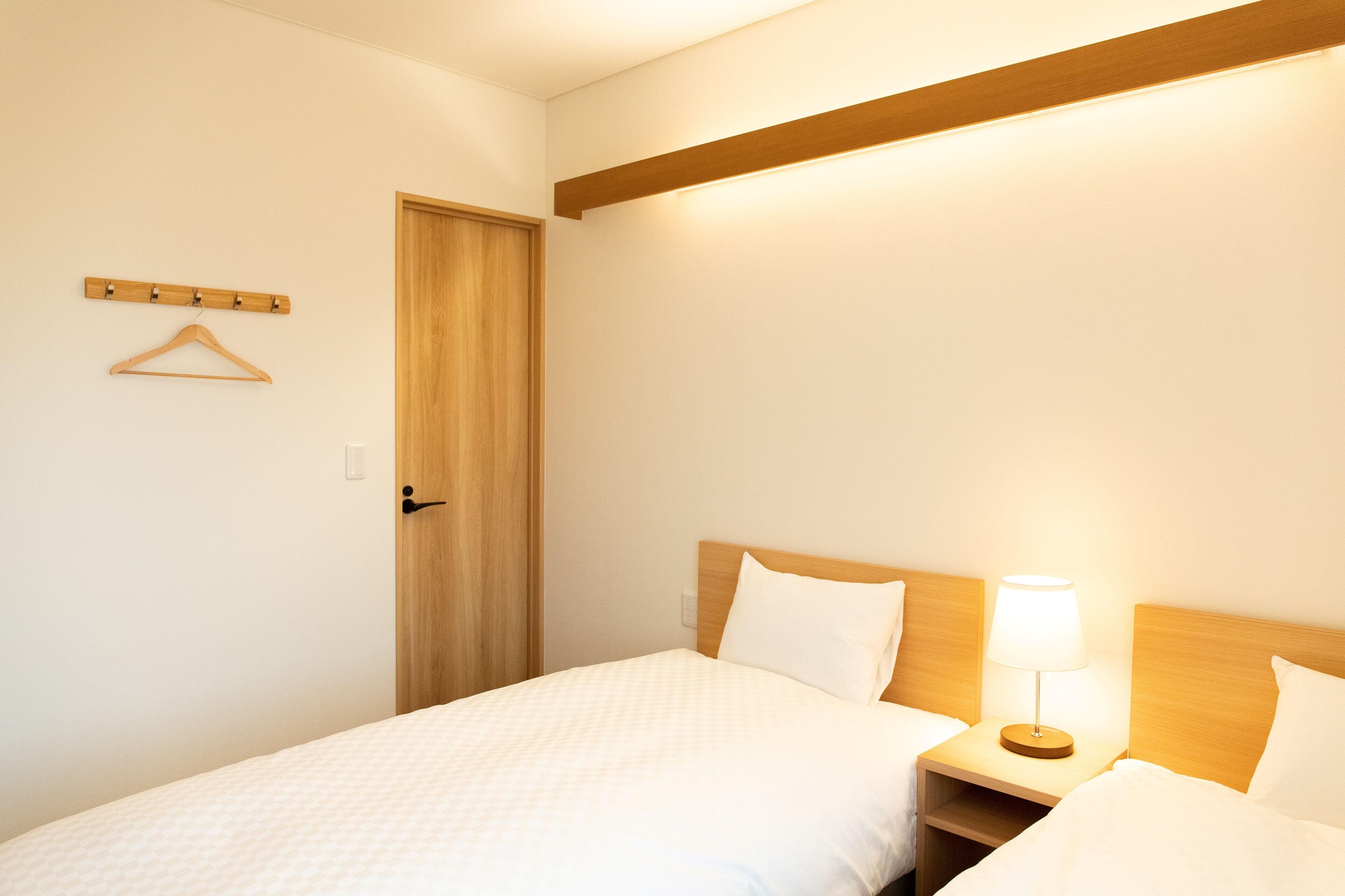 Une chambre d’hôtel avec une porte en bois, un porte-manteau, une table de chevet avec une lampe et deux lits avec du linge de lit blanc.