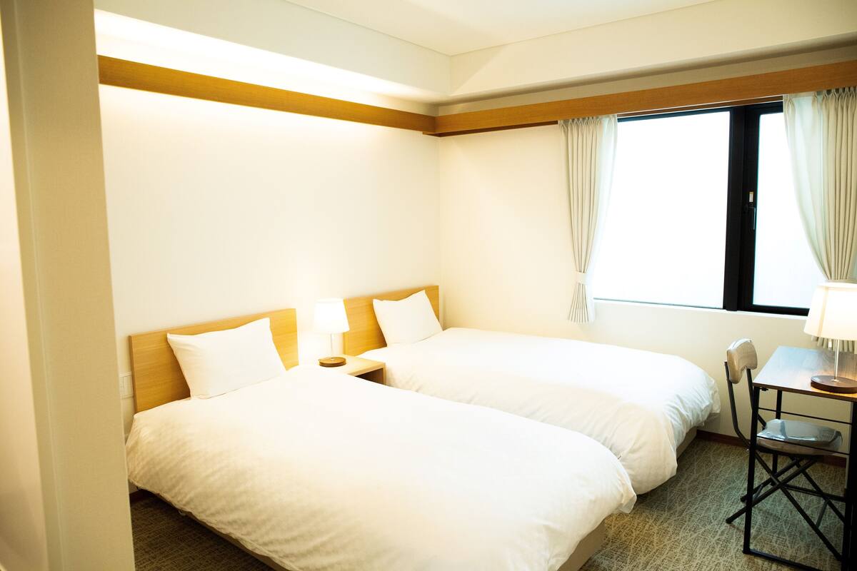 Une chambre d’hôtel avec deux lits, une petite table avec une chaise et une fenêtre avec des rideaux.