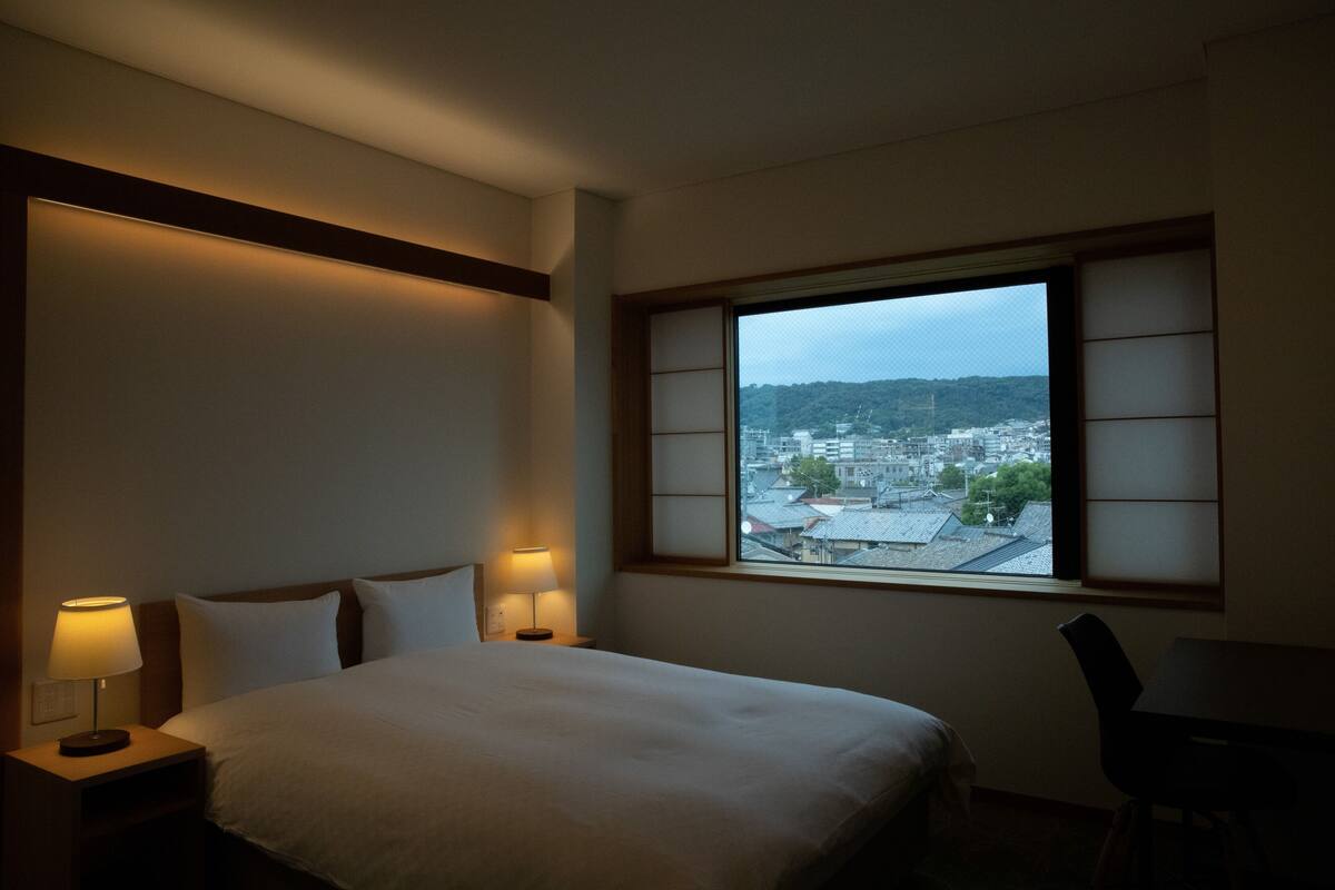 Une chambre d’hôtel avec un lit, deux lampes de chevet, une fenêtre donnant sur un paysage urbain et un fauteuil.