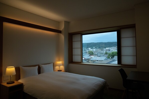 A hotel room with a bed, two bedside lamps, a window with a view of a cityscape, and a chair.