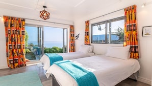 Habitación de hotel con dos camas, ventanas amplias, vista al mar y cortinas coloridas.
