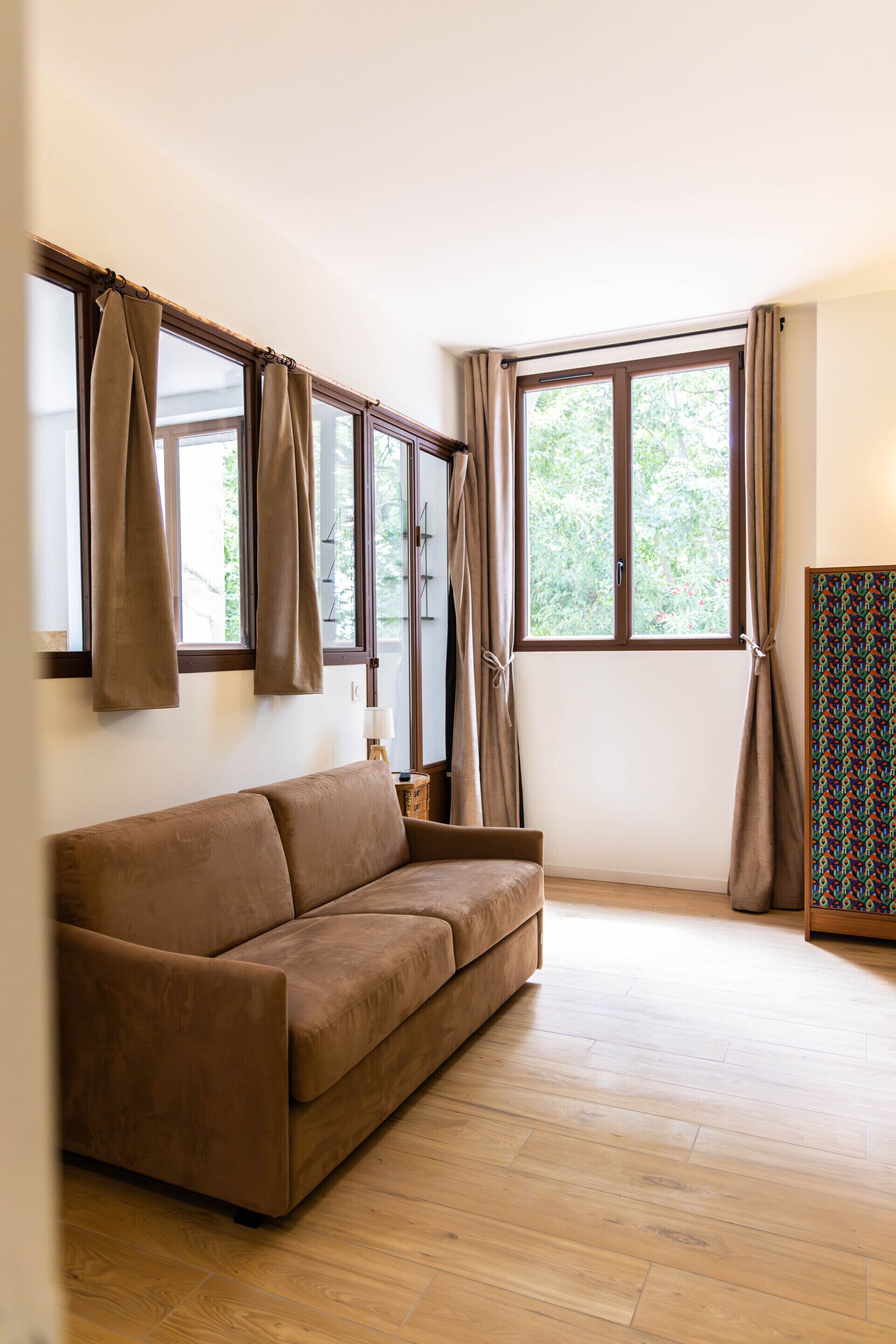 A room with a brown sofa, wooden flooring, and large windows with curtains.