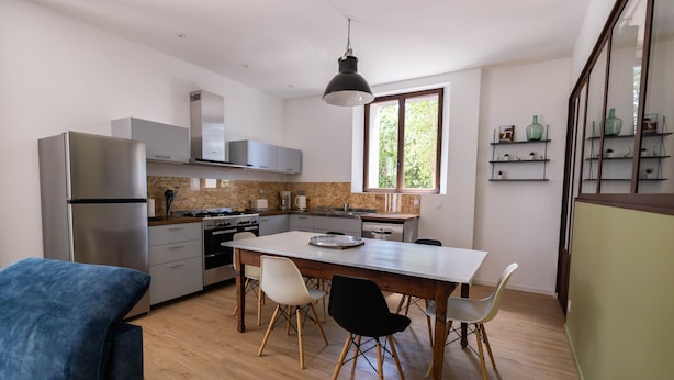 Een moderne keuken met roestvrijstalen apparatuur, een eettafel met stoelen en een groot raam dat zorgt voor veel daglicht.