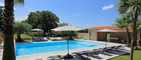 En swimmingpool med liggestole og parasoller, omgivet af palmer og en murstensbygning.