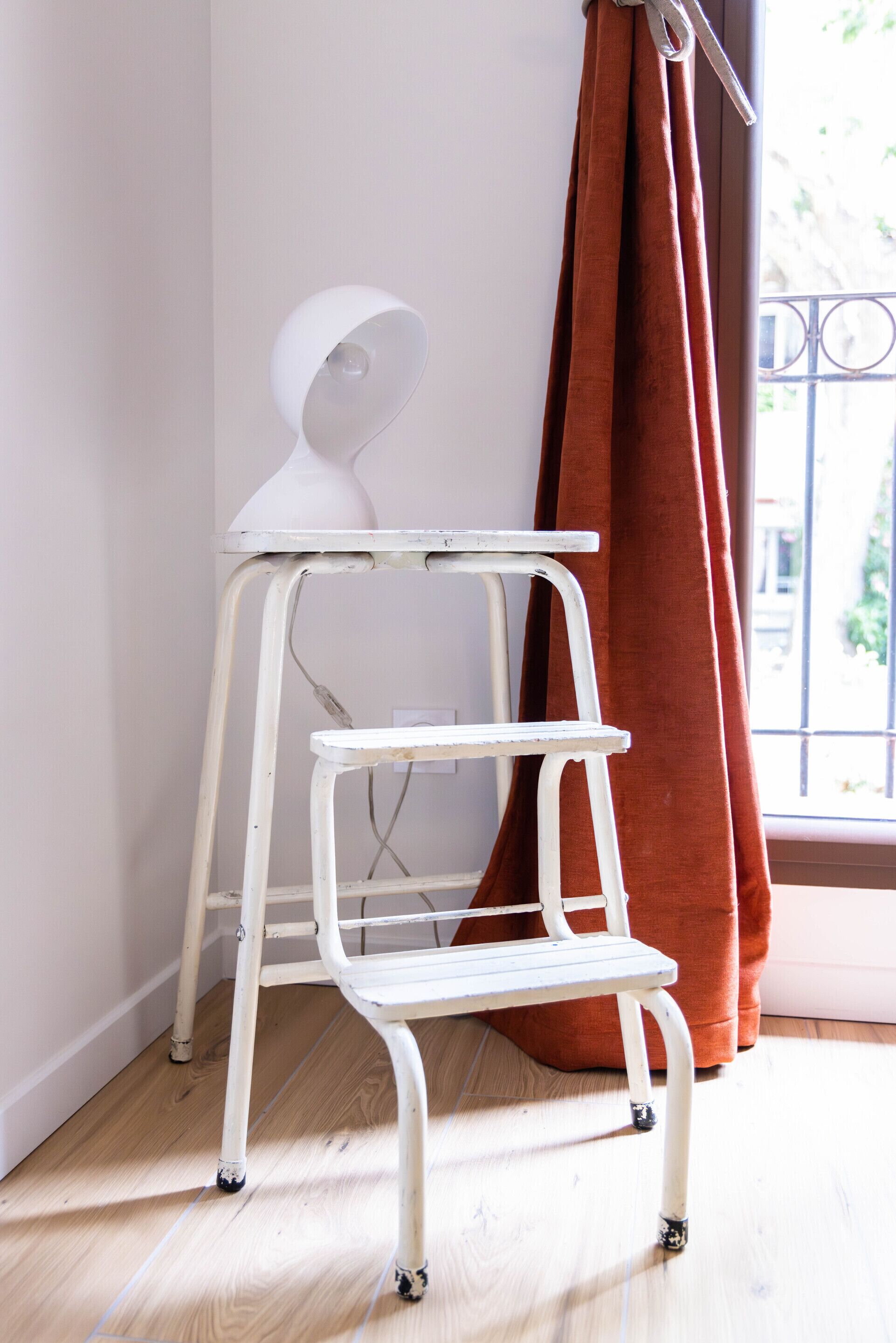 A white metal desk with a curved backrest and a shelf, positioned in a room with a window and a brown curtain.