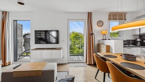 Eine moderne Wohnung mit Wohnzimmer, Essbereich und Küche. Ein Fernseher ist an der Wand montiert, es gibt einen Esstisch mit Stühlen und eine Küche mit Herd und Schränken.