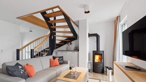 Ein modernes Wohnzimmer mit grauem Sofa, einer Holztreppe und einem Kaminofen.