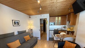 A compact living space with a grey sofa, a black armchair, and a dining table with chairs. A kitchen with wooden cabinets and appliances is visible in the background. - Ski-In/Ski-Out Studio at Les Gets, Close to Village Center (Les Gets)