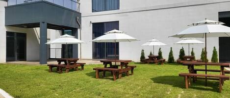 Un patio con mesas y sombrillas al aire libre, rodeado por un edificio con amplias ventanas y un balcón.