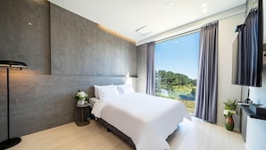 A modern bedroom with a large bed, a nightstand, a lamp, and a view of greenery through the window. - Hillsnorkelbeach hotel (Samcheok)