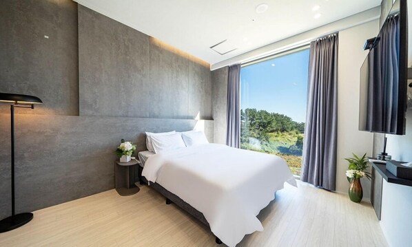 Un dormitorio moderno con una cama grande, una mesita de noche, una lámpara y vistas a la vegetación por la ventana.