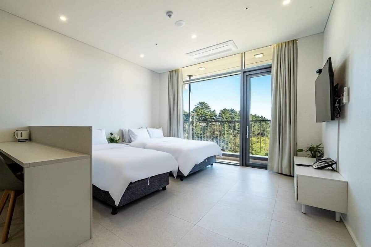 A modern hotel room with a desk, two beds, a balcony with a view, and a TV on the wall.