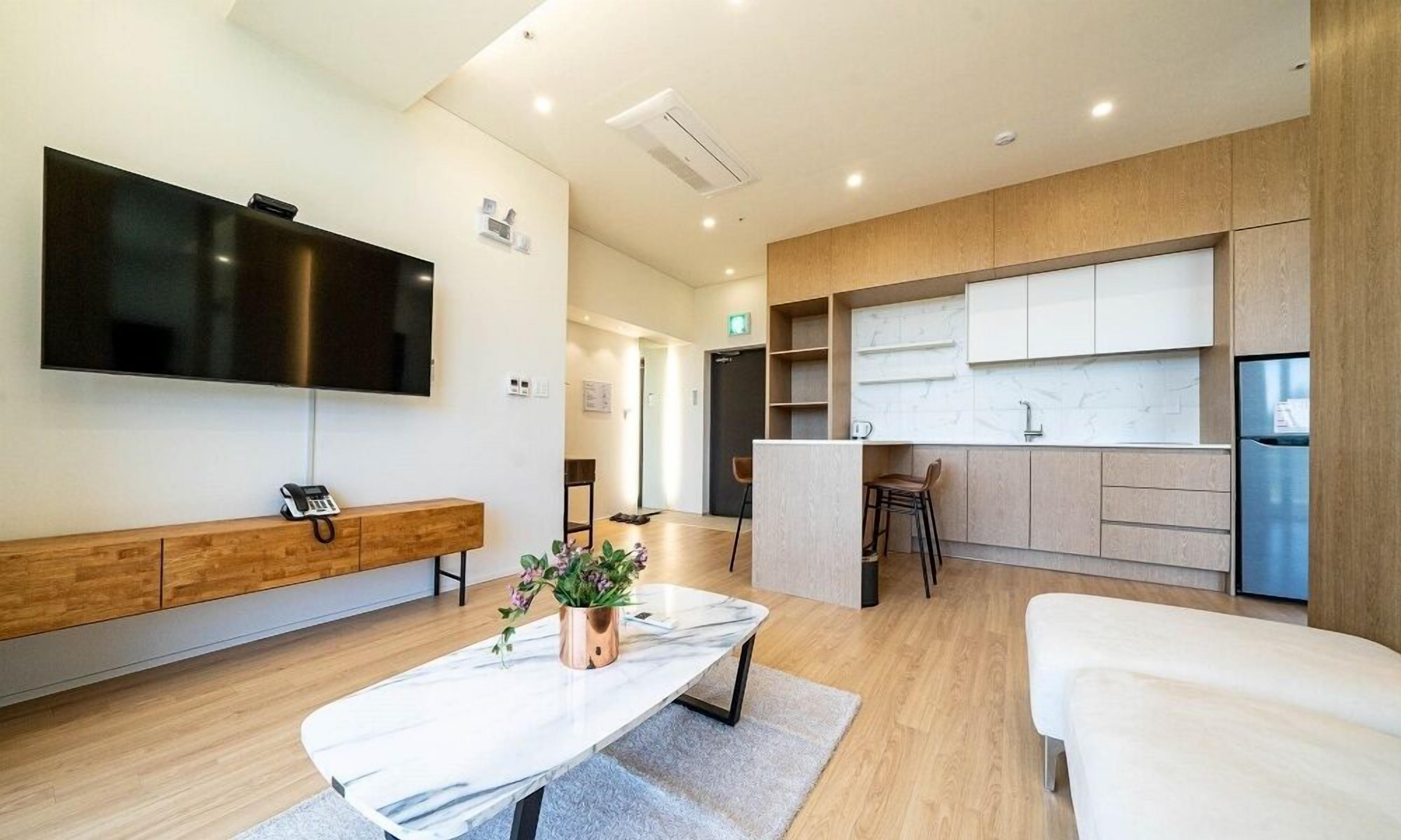 A modern hotel room with a flat-screen TV, a wooden console table, a marble table, and a kitchenette area.