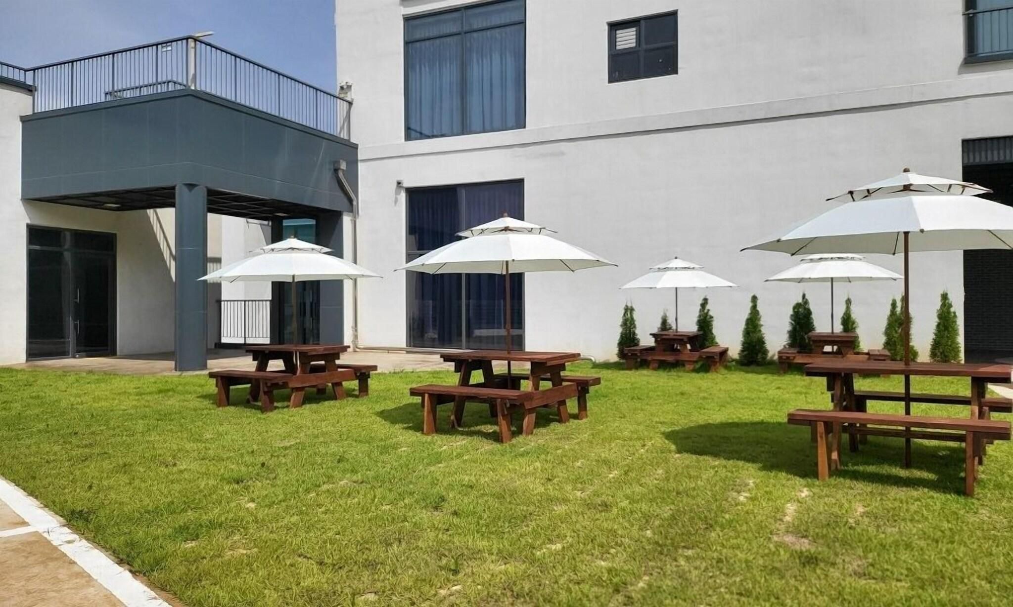 A courtyard with multiple wooden picnic tables and white umbrellas, surrounded by a well-maintained lawn and a modern building with large windows.