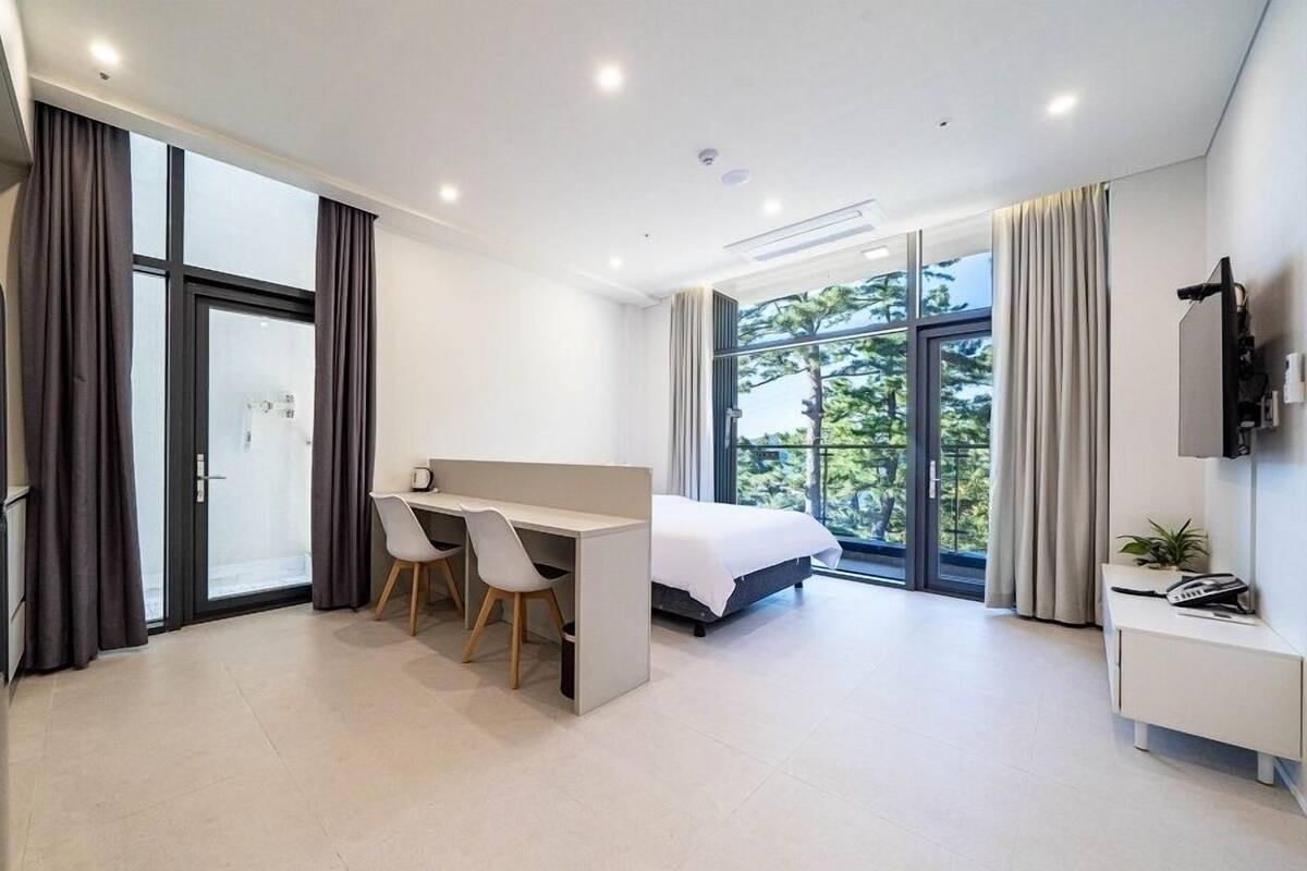 A modern hotel room with a bed, a desk with two chairs, a refrigerator, a TV mounted on the wall, and large windows offering a view of trees.