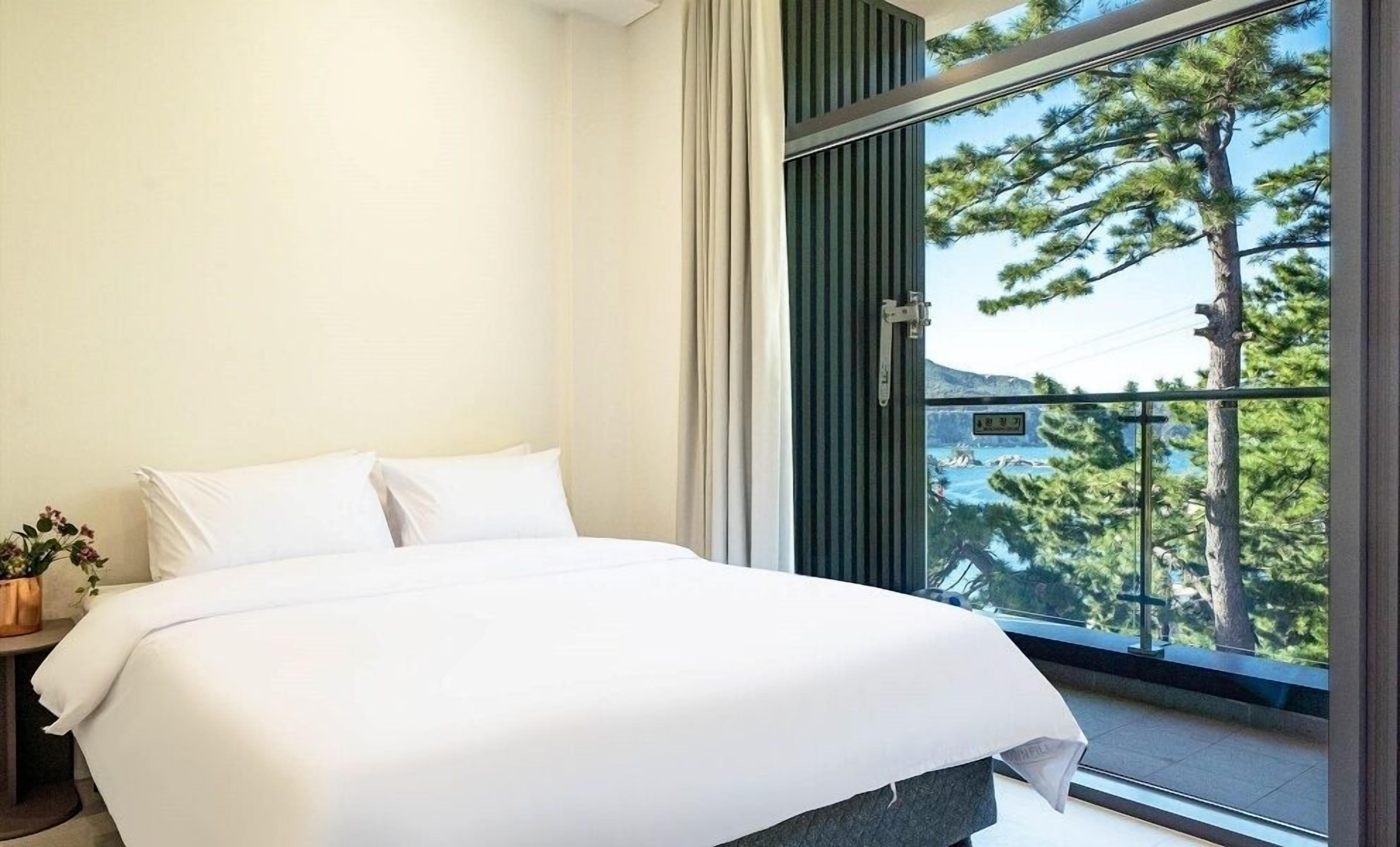 Un dormitorio moderno con una cama grande, una mesita de noche pequeña con una planta y vistas a árboles a través de un ventanal amplio.