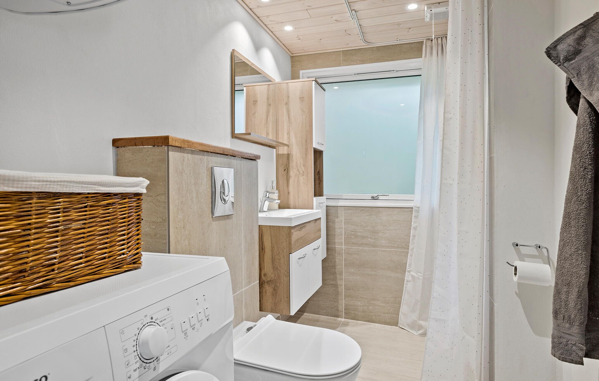 Un baño pequeño con inodoro blanco, una cesta de mimbre a la izquierda, tocador de madera y cabina de ducha con mampara de vidrio.