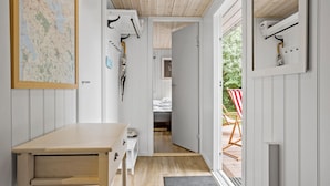 A narrow hallway with a wooden bench, a map on the wall, and a door leading to a room with a bed and a red and white striped chair outside.