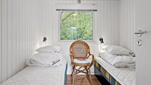 A small, single-room accommodation with two beds, a window with a view of greenery, and a wicker chair.