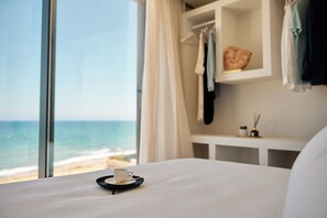 A hotel room with a bed, a small table with a cup and saucer, a shelf with clothes and a basket, and a view of the ocean through large windows. - ANCON SUITES (Carboneras)