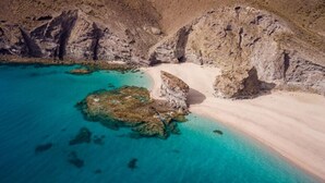 Vista aerea di una zona costiera con acque turchesi cristalline, formazioni rocciose e una spiaggia sabbiosa.