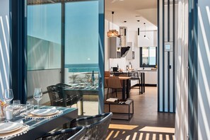 A modern dining area with a view of the ocean, a kitchen in the background, and a balcony with a glass railing. - ANCON SUITES (Carboneras)