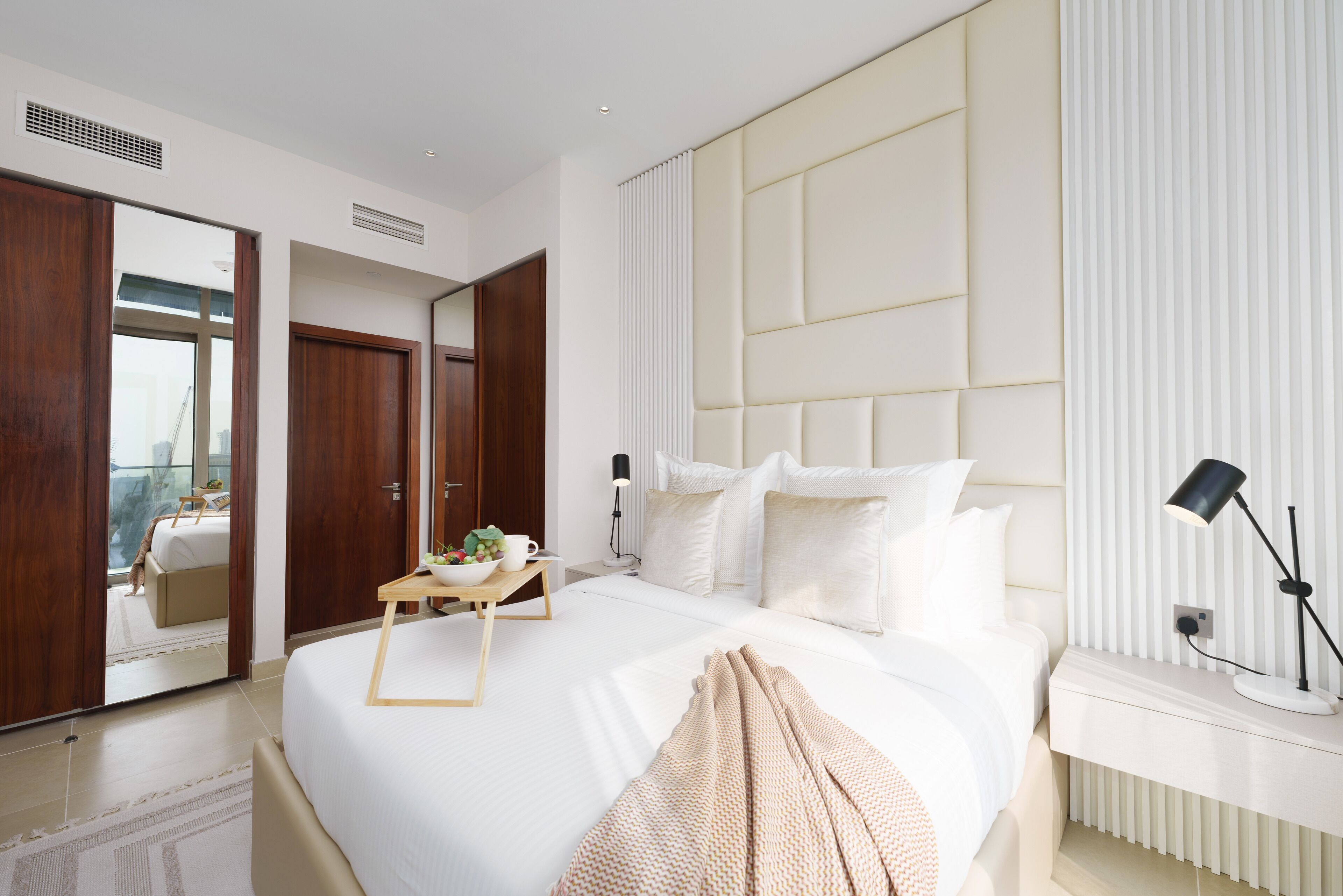 A modern hotel room with a large bed, a bedside table with a lamp, a small table with a bowl of fruit, and a view through an open door.