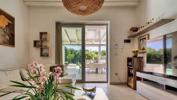 A modern living room with a sofa, coffee table, and TV stand. There is a sliding glass door leading to an outdoor patio. - 3512 Villa Mediterranea by Perle di Puglia (Ostuni)