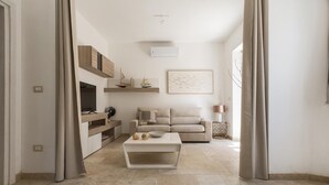 A modern living room with a sofa, coffee table, and decorative items. There is a wooden shelf with small decorative objects and a framed picture on the wall. - 3512 Villa Mediterranea by Perle di Puglia (Ostuni)