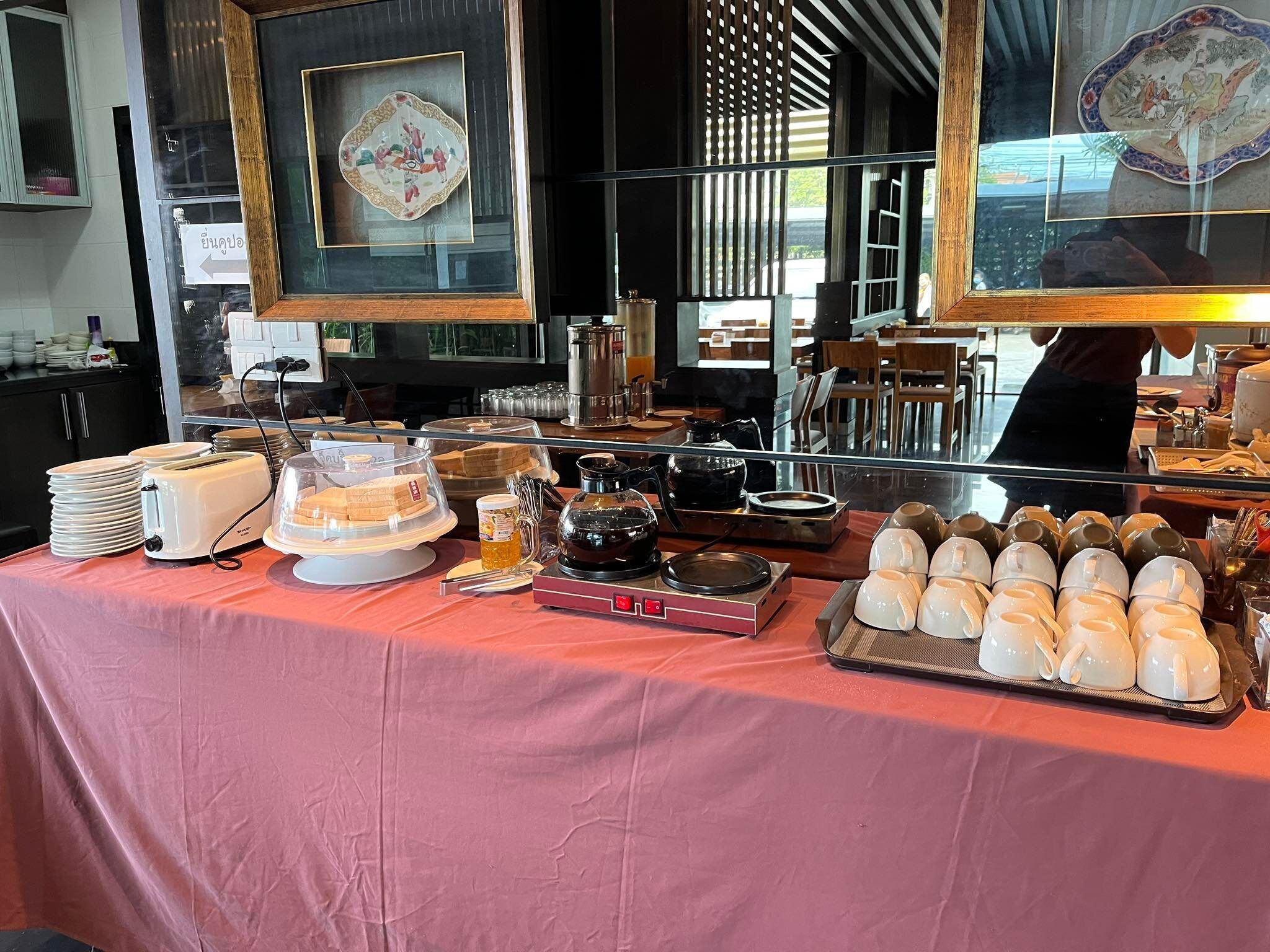 Disposição de buffet com diversos alimentos, uma máquina de café e um chaleira sobre uma toalha de mesa rosa.
