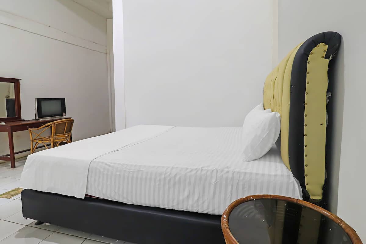 A neatly made bed with a white bedspread and pillows, a wooden nightstand with a television, and a chair in a room with white walls and a tiled floor.