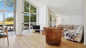 Una habitación amplia con un gran ventanal corredera que da a un espacio exterior, un sillón de cuero, un sofá y un televisor sobre un mueble.