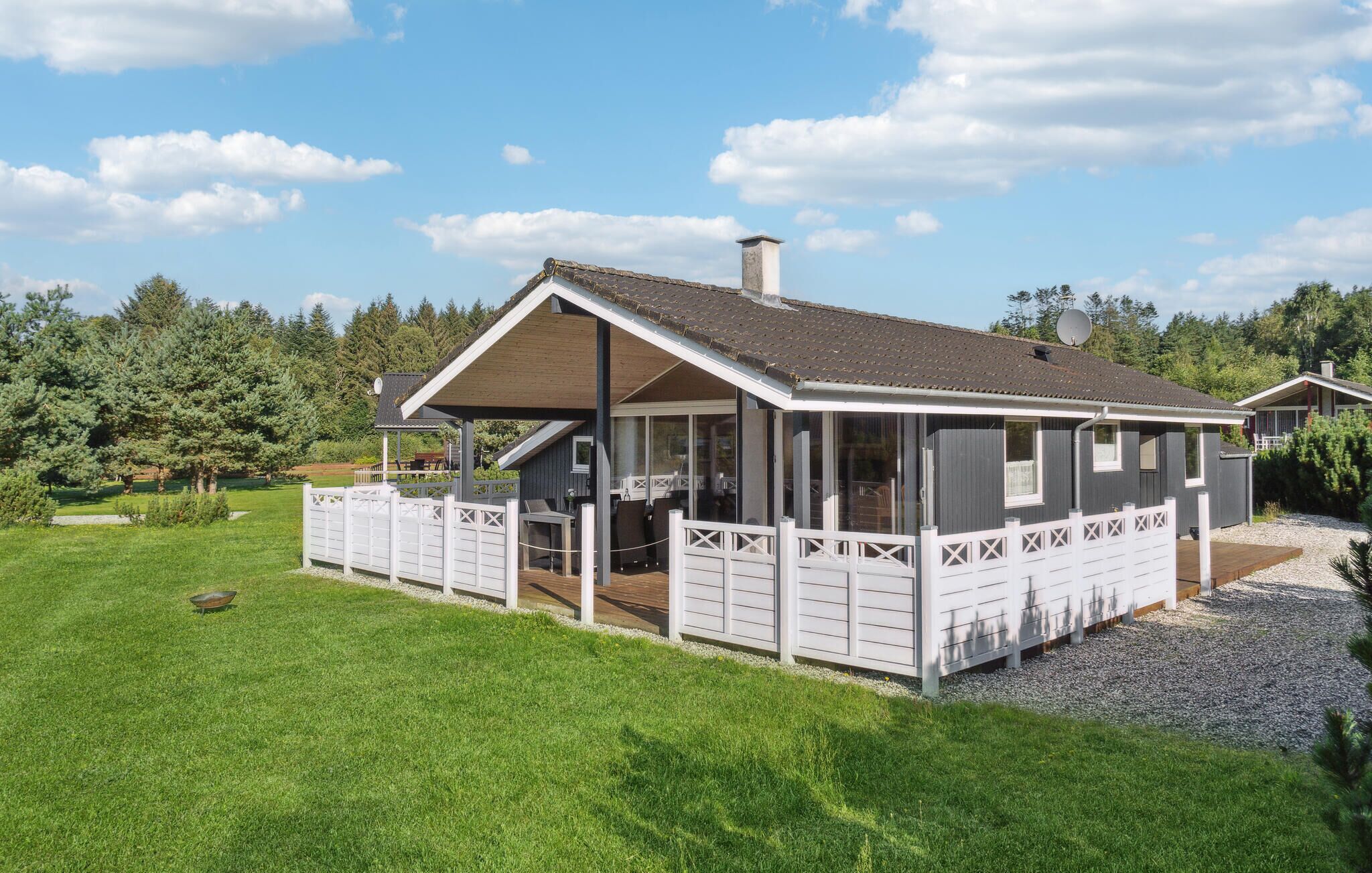 A single-story cabin with a covered patio, surrounded by a white picket fence, set in a grassy area with trees in the background.