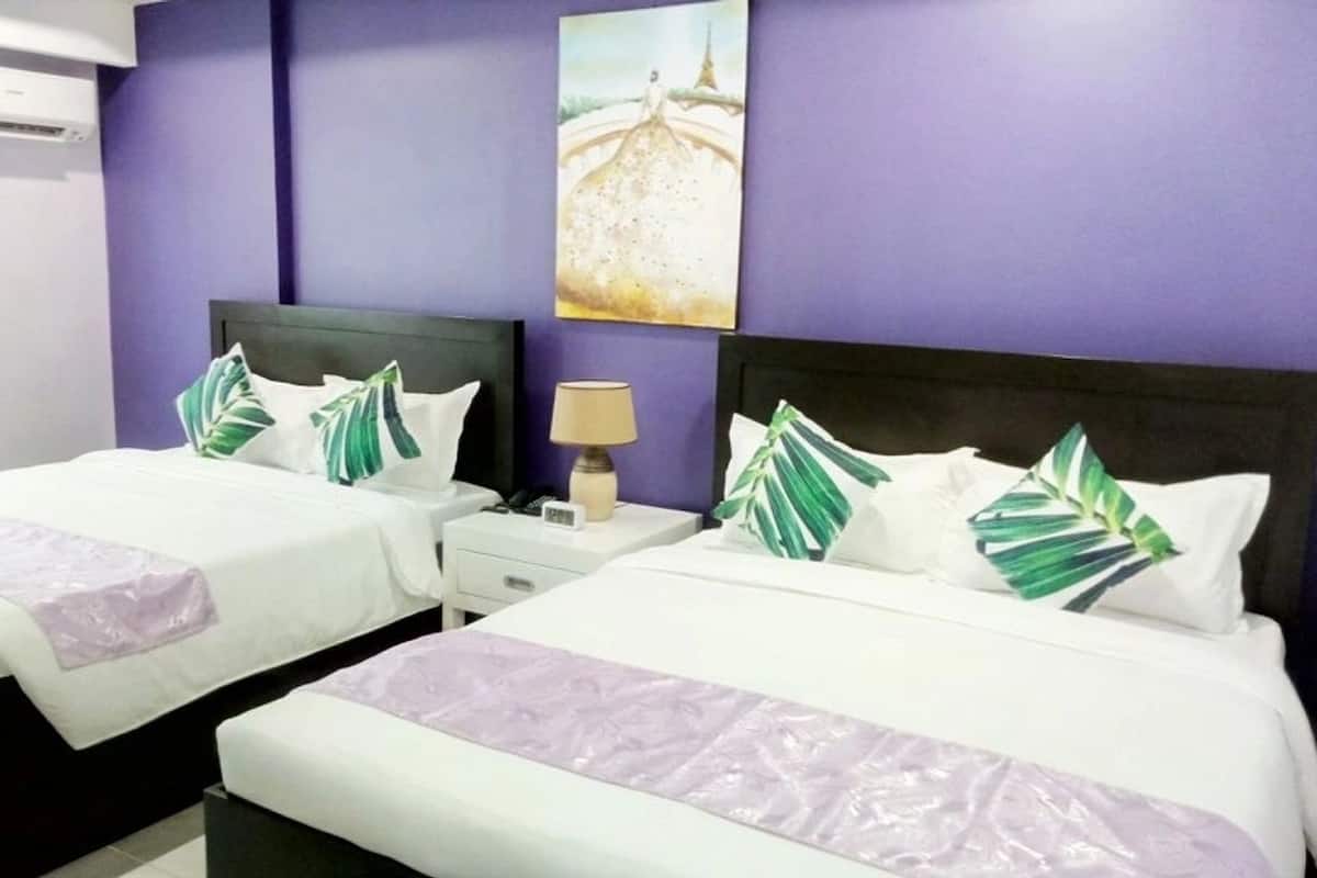 A hotel room with two beds, each with white bedding and green leaf pillows. There is a nightstand with a lamp between the beds, and a painting on the wall above the beds.