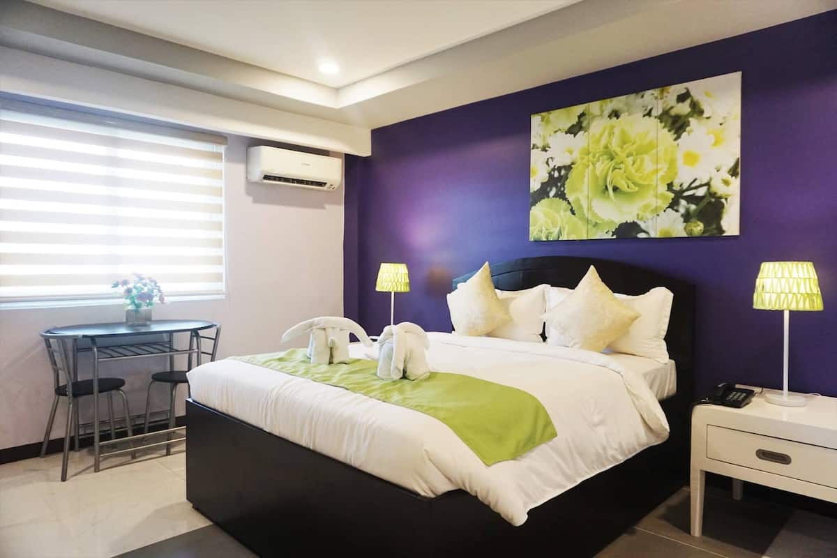 A modern hotel room with a large bed, two bedside lamps, a small table with chairs, and a window with blinds.