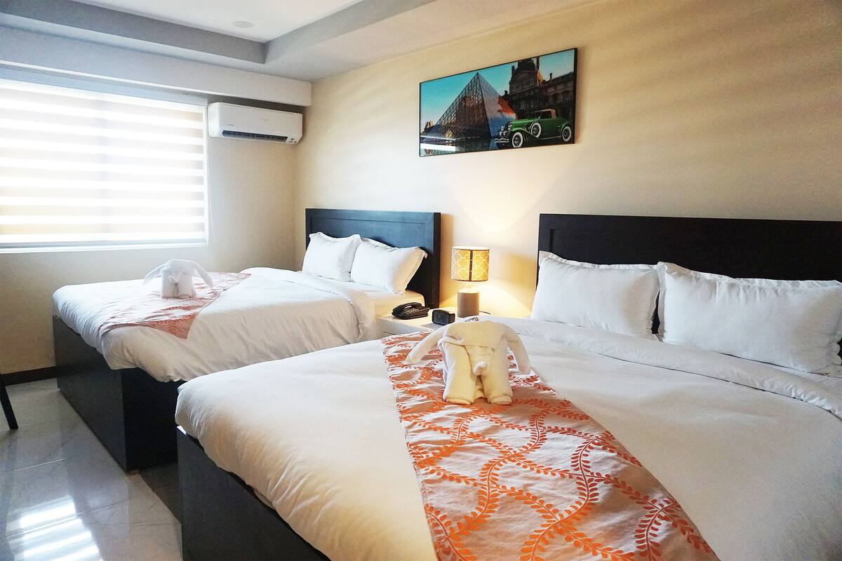 A hotel room with two beds, a painting on the wall, a lamp, and an air conditioning unit.