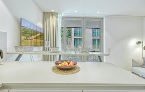 A modern kitchen with a white countertop, a bowl of fruit, and a dining area with a table and chairs.
