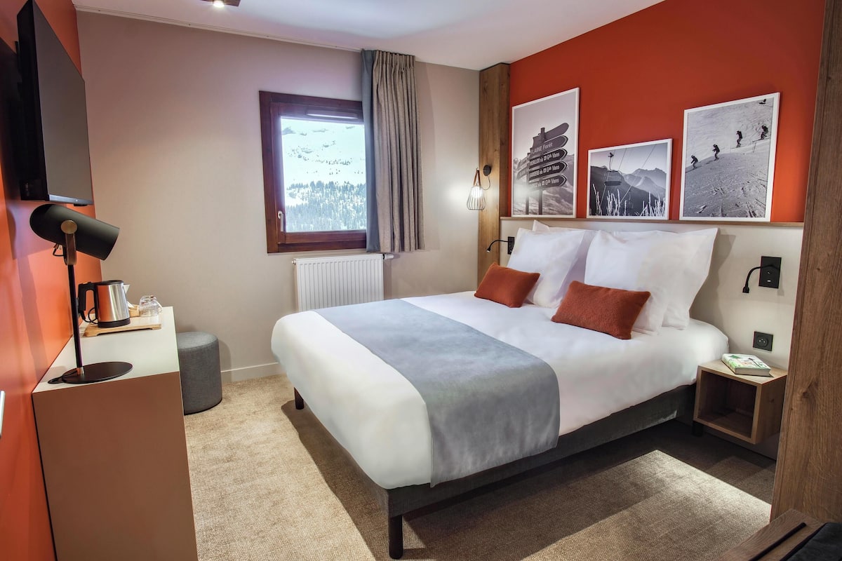 Una habitación de hotel moderna con una cama grande, una cafetera, una mesita de noche y una ventana con vistas a un paisaje nevado.