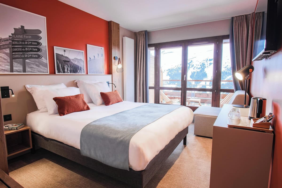 Habitación de hotel con una cama grande, un escritorio con lámpara y una ventana con vistas a las montañas.