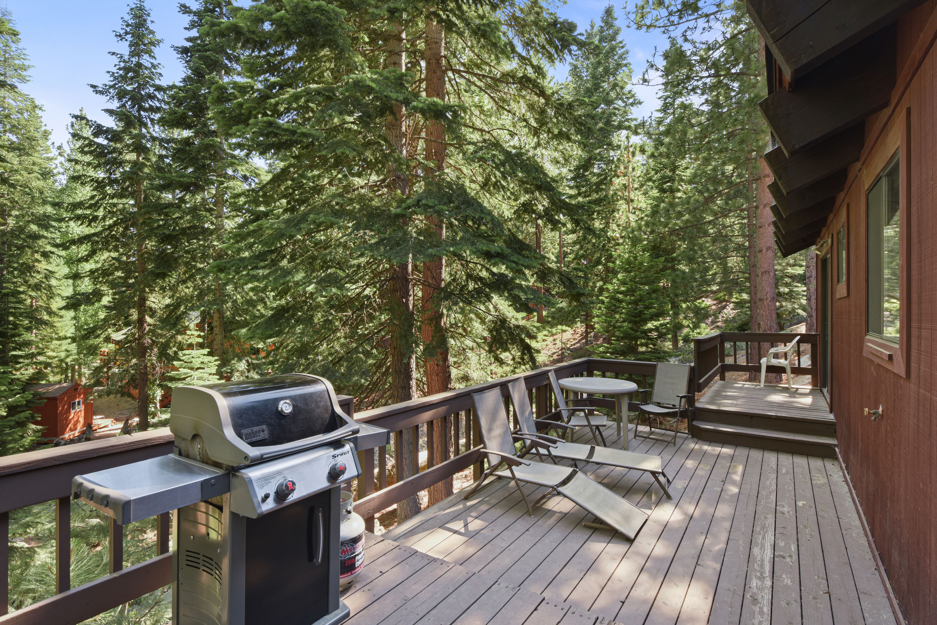 A deck with a grill, chairs, and a table, surrounded by tall trees.