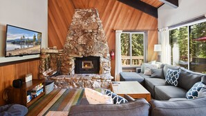 Ein gemütliches Wohnzimmer mit Kamin aus Stein, Holzbalkendecke, einer bequemen Couch und einem großen Fenster mit Blick ins Freie.