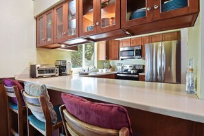 A kitchen with wooden cabinets, stainless steel appliances, a bar area with chairs, and a view of greenery outside.