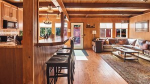 Amplia sala de estar con piso de madera, un conjunto de sofás y una mesa de centro. Hay una cocina con gabinetes de madera y un microondas.