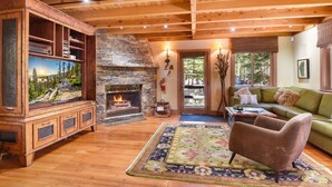 Un salon chaleureux avec une cheminée en pierre, des poutres apparentes en bois au plafond, un canapé vert et un téléviseur posé sur un meuble en bois.