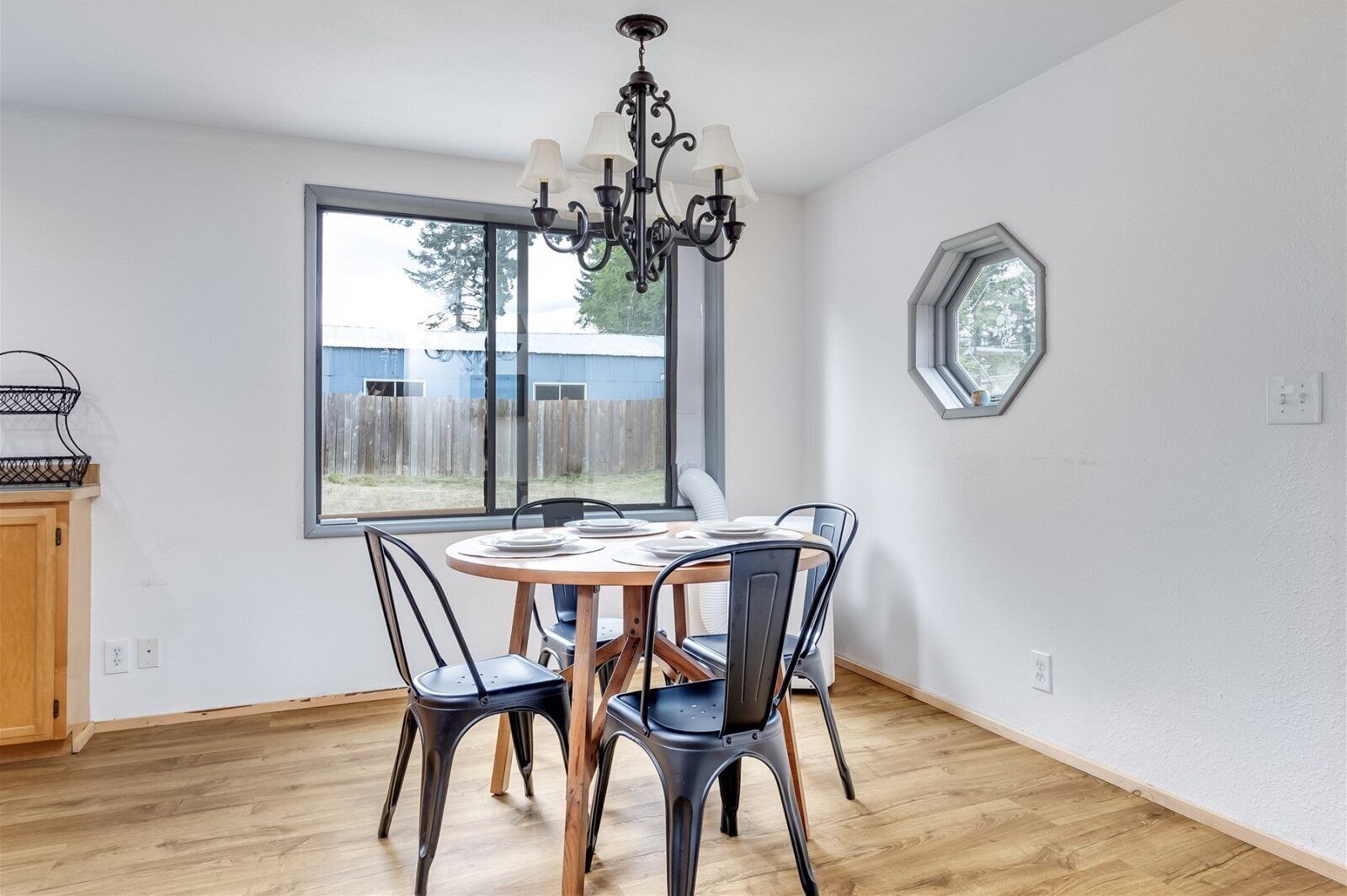 A dining area with a round table, four chairs, a chandelier, and a large window with a view of a wooden fence and trees.