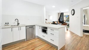 Una cocina moderna con armarios blancos, un lavavajillas de acero inoxidable y un microondas. Se ve un piso de madera y un comedor al fondo.