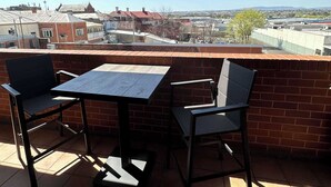 Un balcón con una mesa y sillas, un muro de ladrillo y vistas a edificios y colinas.